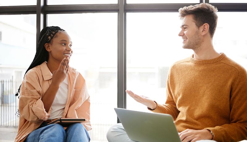 Two students, holding a tablet and a laptop, sit next to a window and discus assistive technology