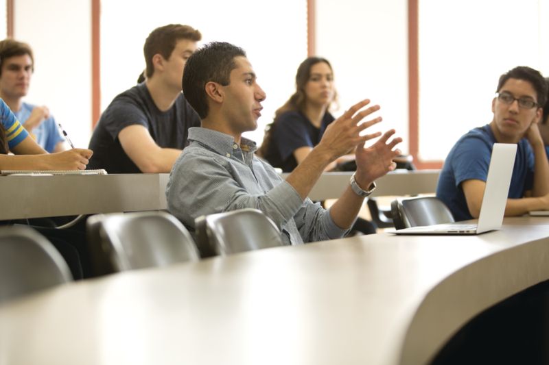 A student gestures while speaking during a college classroom discussion as classmates listen from tiered seating, with an open laptop on the desk nearby