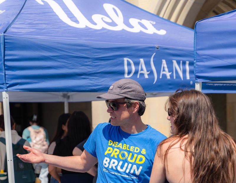 A UCLA Center for Accessible Education staff member wearing a “Disabled & Proud Bruin” T-shirt speaks with a student at an outdoor campus resource fair beneath blue UCLA canopy.