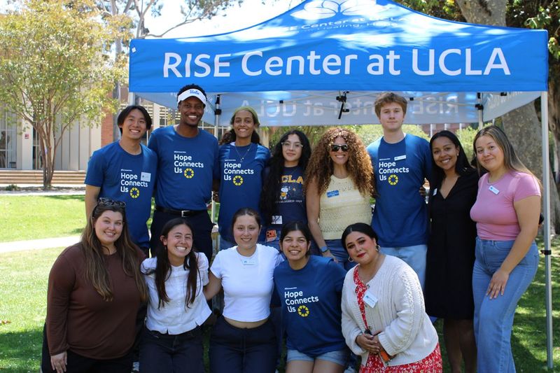 Group of 13 UCLA students and staff pose outdoors beneath a blue “RISE Center at UCLA” canopy. Several wear blue “Hope Connects Us” T-shirts, while others wear casual clothing. Trees, grass, and campus buildings are visible in the background
