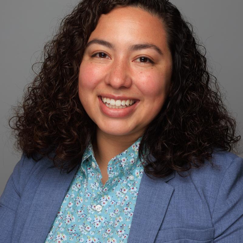 Headshot of woman with brown and long curly hair wearing a blue patterned shirt and blazer